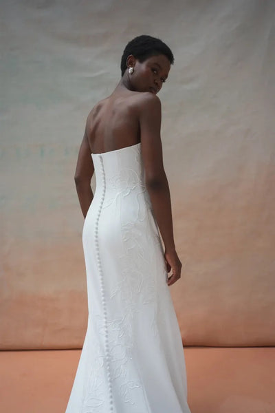 A person wearing the Tricia Wedding Dress by Jenny Yoo from Bergamot Bridal, a strapless white gown adorned with floral organza applique, stands with their back facing the camera. The photo background features a gradient of warm colors.