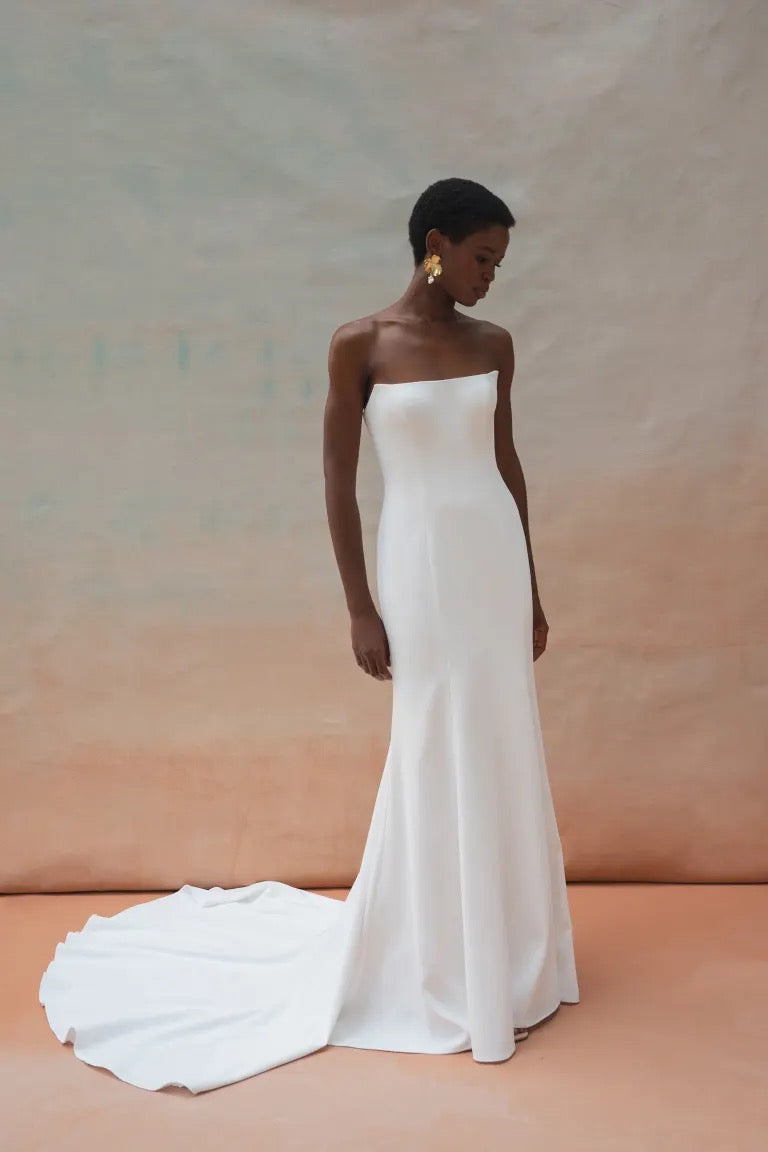 A woman stands wearing the strapless white Tricia Wedding Dress by Jenny Yoo from Bergamot Bridal, which features a floor-length train and floral organza appliques, against a neutral background. She looks downwards, her short hair perfectly complementing the elegant fit-and-flare silhouette. Dangling earrings add a touch of sparkle to her ensemble.