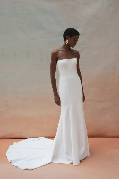 A woman stands wearing the strapless white Tricia Wedding Dress by Jenny Yoo from Bergamot Bridal, which features a floor-length train and floral organza appliques, against a neutral background. She looks downwards, her short hair perfectly complementing the elegant fit-and-flare silhouette. Dangling earrings add a touch of sparkle to her ensemble.