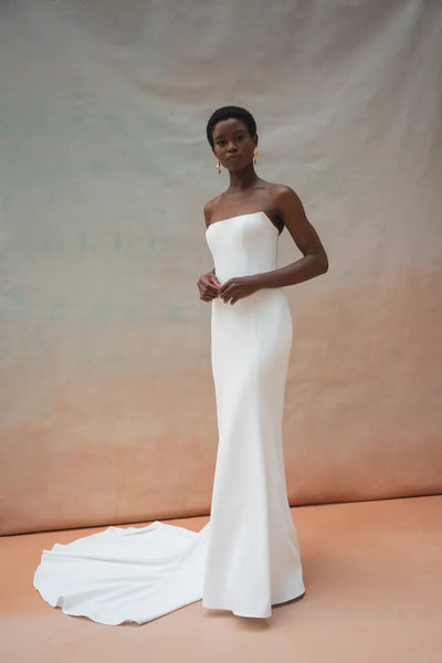 A woman poses in a strapless white Tricia Wedding Dress by Jenny Yoo for Bergamot Bridal, showcasing a fit and flare silhouette with a long train and intricate floral organza appliqué, all set against a neutral backdrop.