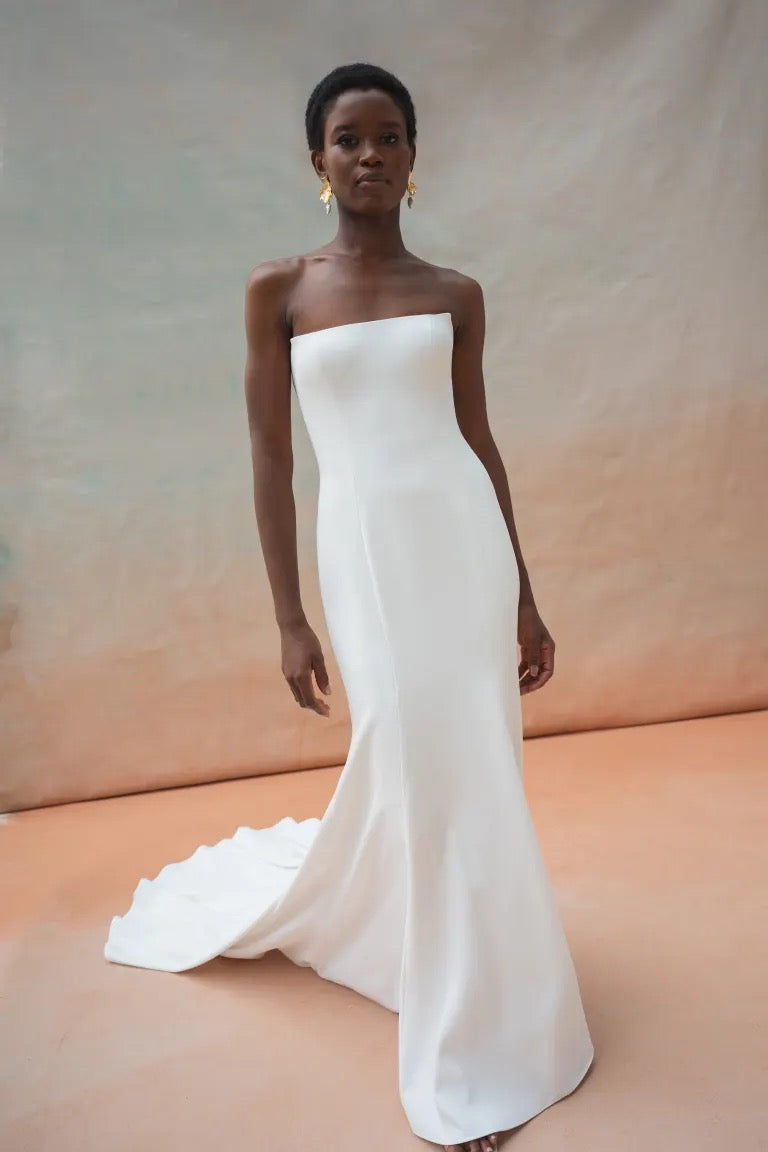 A person wearing a strapless Tricia Wedding Dress by Jenny Yoo from Bergamot Bridal, featuring a small train and delicate floral organza applique, standing against a neutral-colored backdrop.