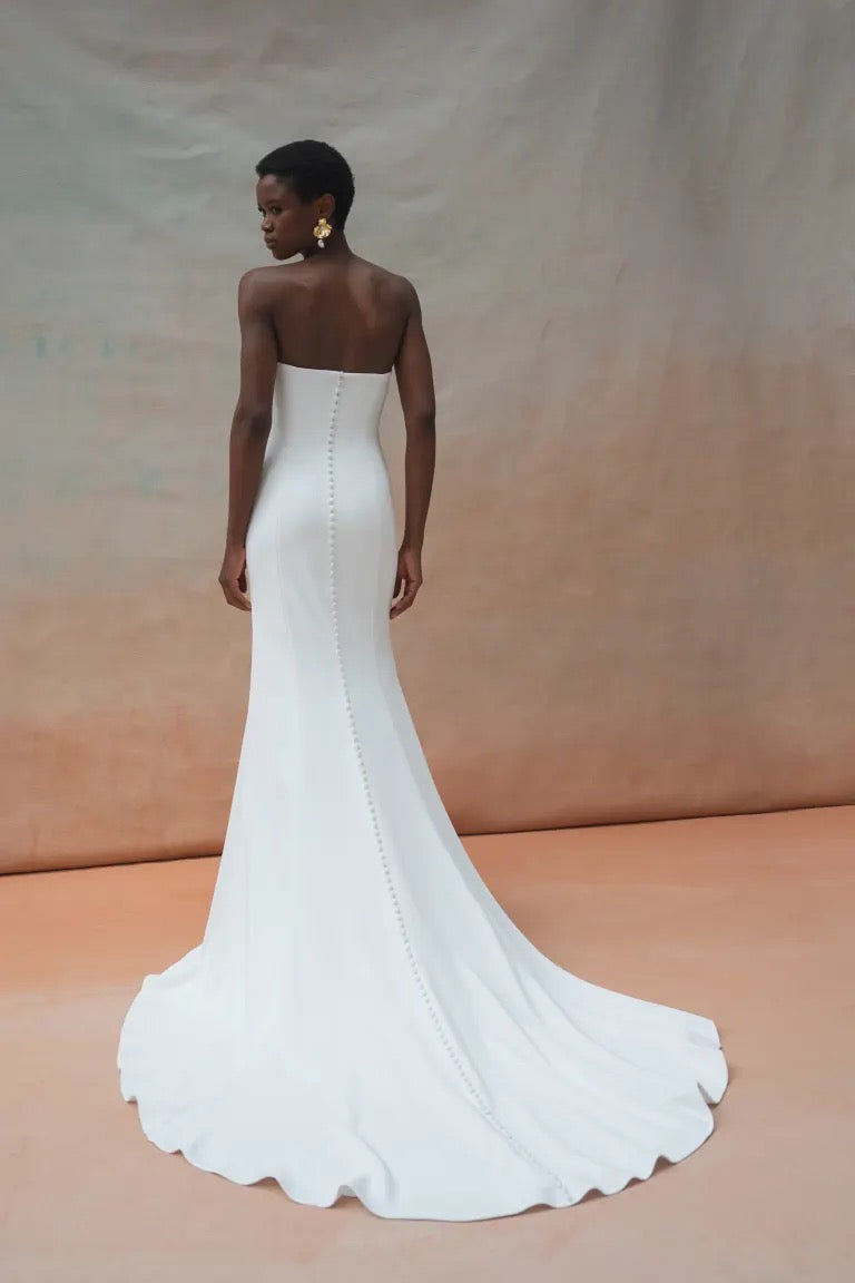 A woman in a strapless white Tricia Wedding Dress by Jenny Yoo from Bergamot Bridal stands with her back to the camera, showcasing a fit and flare silhouette, long train, and button details along the back.