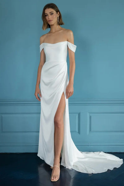 Woman posing in an elegant off-the-shoulder Bergamot Bridal Jenny Yoo Wedding Dress with a thigh-high slit against a blue wall.