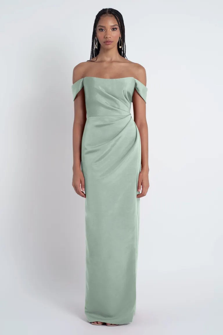 Woman in an elegant, Jenny Yoo bridesmaid dress with off-the-shoulder sleeves, crafted from luxe Faille fabric, in sage green standing against a white background from Bergamot Bridal.