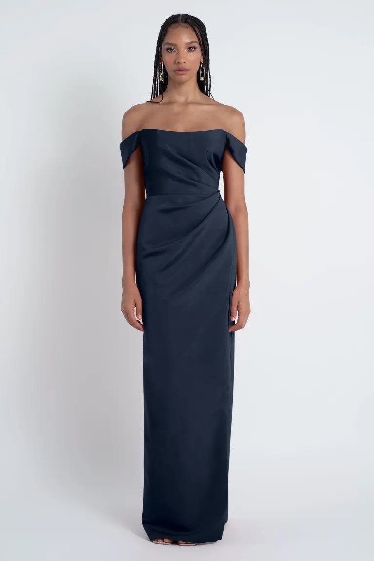 A woman in an off-the-shoulder navy blue Jenny Yoo Bridesmaid Dress by Waverly standing against a white background from Bergamot Bridal.