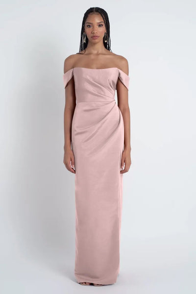 Woman in a Luxe Failile fabric off-the-shoulder blush pink Jenny Yoo Bridesmaid Dress standing against a white background from Bergamot Bridal.