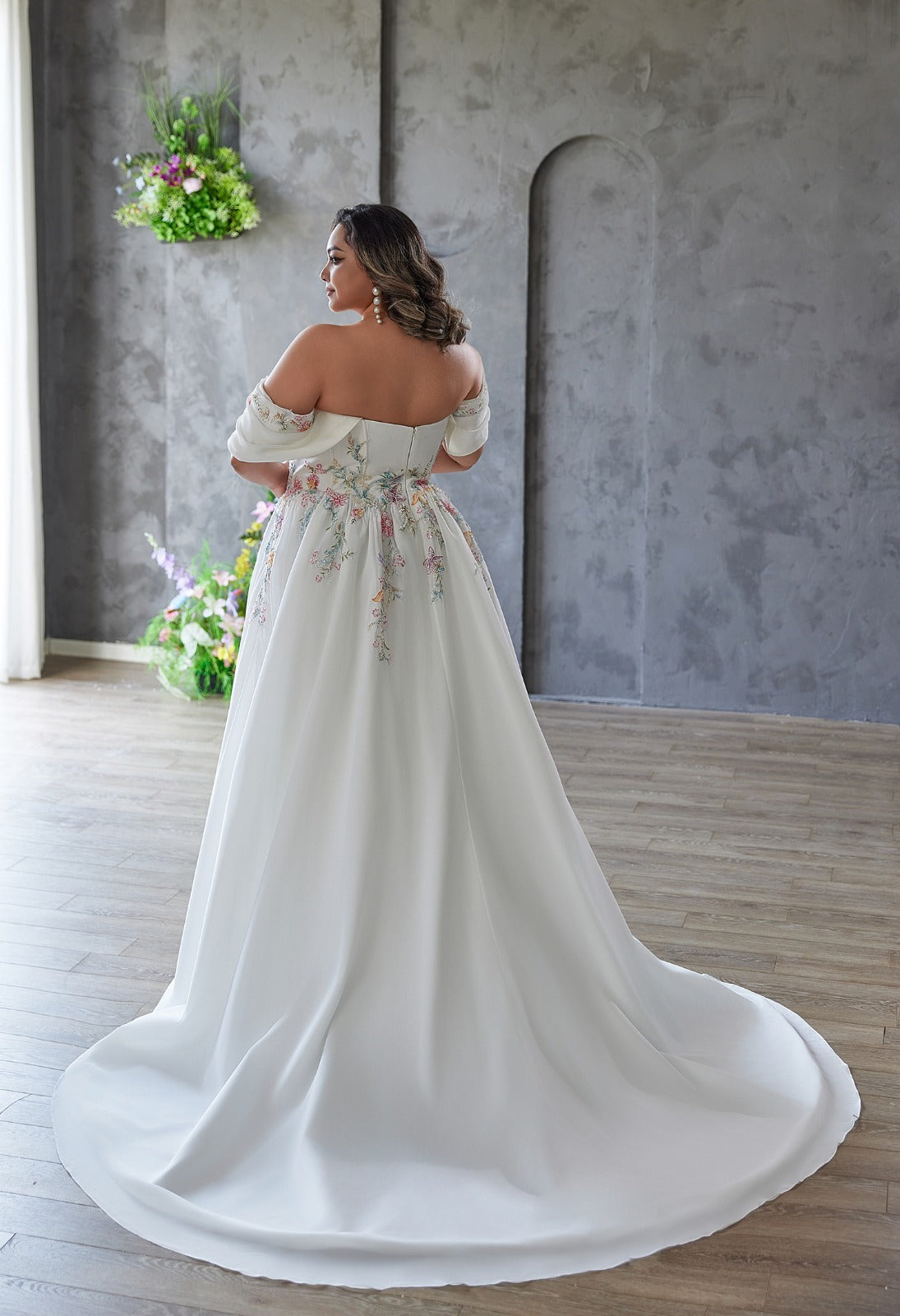 A woman in a Bergamot Bridal Coloured Embroidered A-Line Gown with detachable draped sleeves stands indoors on a wooden floor, facing away from the camera.