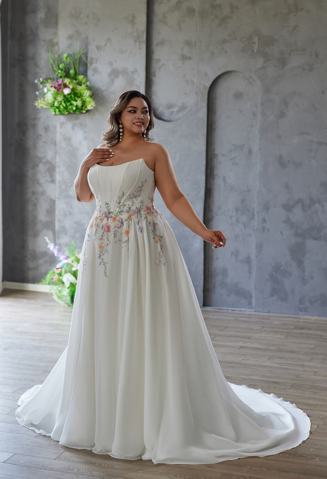 A woman wears the Coloured Embroidered A-Line Gown by Bergamot Bridal, featuring a strapless floral-embroidered bodice and flowing chiffon skirt, standing indoors on a wooden floor with a gray textured wall and hanging plants behind her.