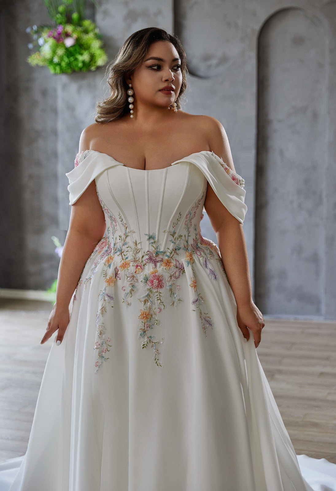 A woman wears the Coloured Embroidered A-Line Gown by Bergamot Bridal, featuring an off-the-shoulder cat eye scoop neckline and floral embroidery, as she stands indoors against a gray textured wall.