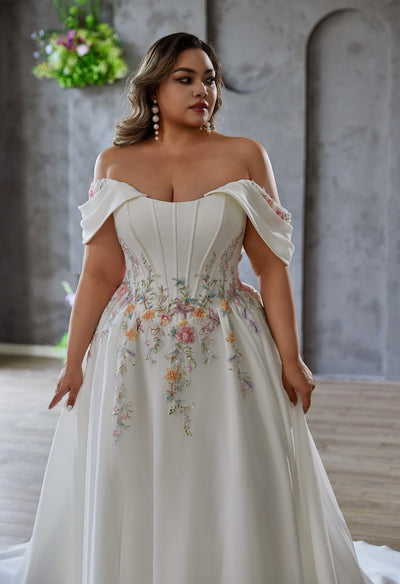 A woman wears the Coloured Embroidered A-Line Gown by Bergamot Bridal, featuring an off-the-shoulder cat eye scoop neckline and floral embroidery, as she stands indoors against a gray textured wall.