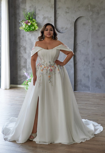A woman wearing the Bergamot Bridal Coloured Embroidered A-Line Gown stands indoors on a wooden floor before a gray textured wall. The dress features an off-the-shoulder design, cat eye scoop neckline, and floral embroidery.