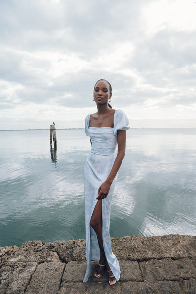 Woman in a printed satin Jenny Yoo Bridesmaid Dress standing by the sea with clouds overhead.