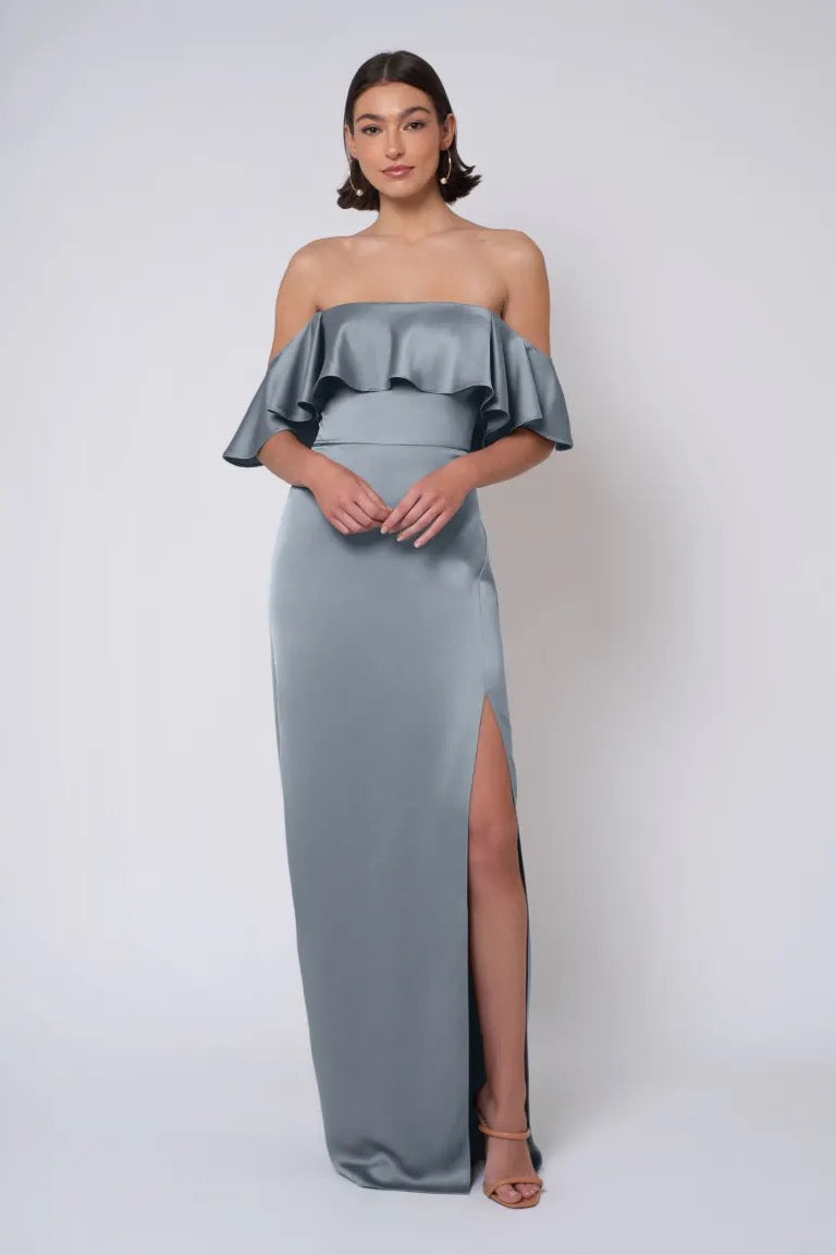 A woman is standing against a plain background, wearing the Winter - Jenny Yoo Bridesmaid Dress by Bergamot Bridal, which features an off-the-shoulder design in blue satin with a thigh-high slit and strapless neckline.