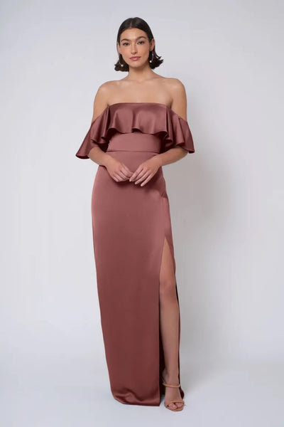 A model wearing the "Winter - Jenny Yoo Bridesmaid Dress" by Bergamot Bridal, featuring an off-the-shoulder, ruffled satin design with a thigh-high slit and strapless neckline, stands against a plain white background.