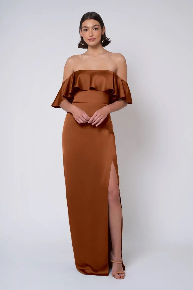 A person wearing the "Winter - Jenny Yoo" Bridesmaid Dress by Bergamot Bridal, an off-the-shoulder, rust-colored gown with ruffle detail and a thigh-high slit, featuring a column skirt, is standing against a plain white background.