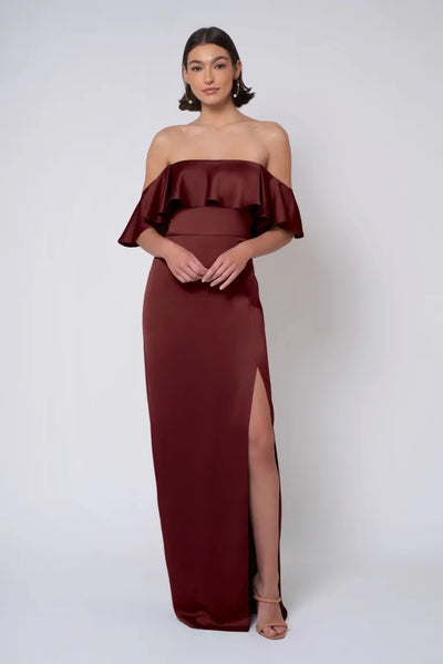 A woman stands against a plain background wearing the Bergamot Bridal Winter - Jenny Yoo Bridesmaid Dress, an off-the-shoulder maroon satin gown featuring a ruffle detail and a thigh-high slit, paired with strappy heels.