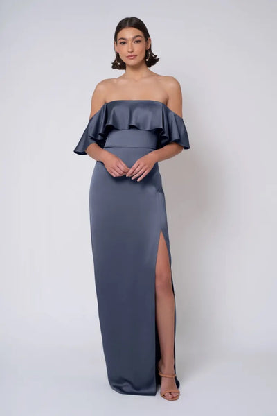 A woman stands in the Winter - Jenny Yoo Bridesmaid Dress by Bergamot Bridal, a strapless dark gray satin gown featuring a ruffled overlay and a thigh-high slit, against a plain white background.