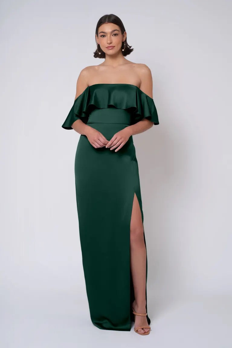 The person is wearing the Winter - Jenny Yoo Bridesmaid Dress by Bergamot Bridal, an off-the-shoulder, dark green, floor-length satin dress featuring a ruffled top and a high slit on the left side; standing against a plain backdrop.