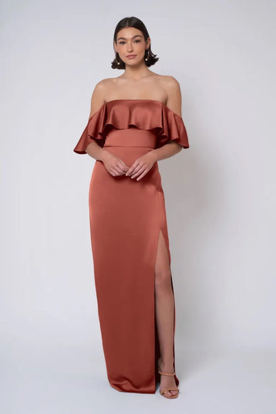 A model showcasing the "Winter - Jenny Yoo Bridesmaid Dress" by Bergamot Bridal, featuring an off-the-shoulder design in rust-colored satin with a ruffled neckline and a column skirt, posed against a plain white background.