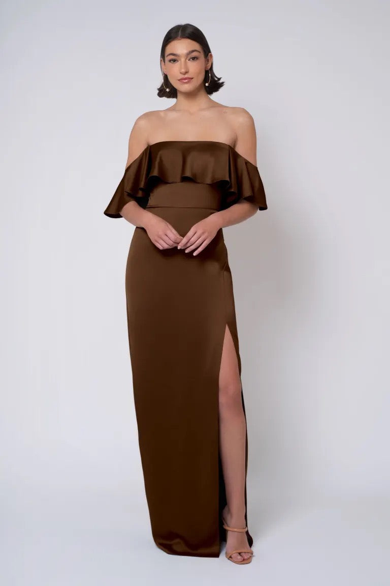 A woman stands against a plain background wearing the Winter - Jenny Yoo Bridesmaid Dress by Bergamot Bridal, featuring a satin finish with ruffled detail and a thigh-high slit. She has shoulder-length hair, holds her hands in front of her, and the strapless neckline accentuates the sophisticated look.