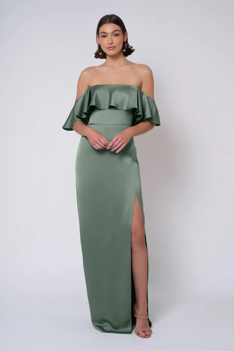 A woman stands against a plain background wearing the Winter - Jenny Yoo Bridesmaid Dress by Bergamot Bridal in deep green, featuring a thigh-high slit. The elegant gown showcases an off-shoulder, strapless neckline and column skirt design, perfectly complementing her short hair.