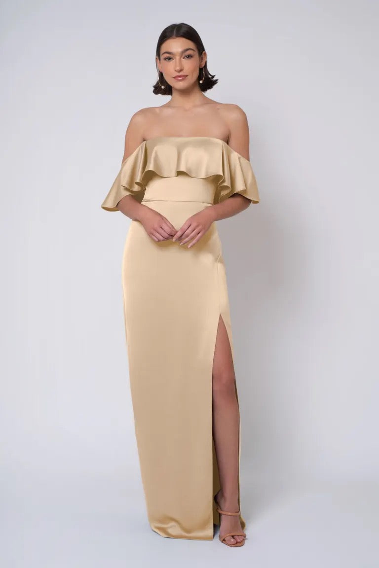 A woman models an elegant Jenny Yoo Bridesmaid Dress off-the-shoulder gold gown with a thigh-high slit from Bergamot Bridal.