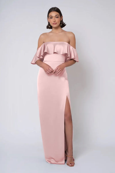 A woman in an elegant Jenny Yoo Bridesmaid Dress from Bergamot Bridal satin pink off-shoulder gown with a strapless neckline and a thigh-high slit posing against a light background.