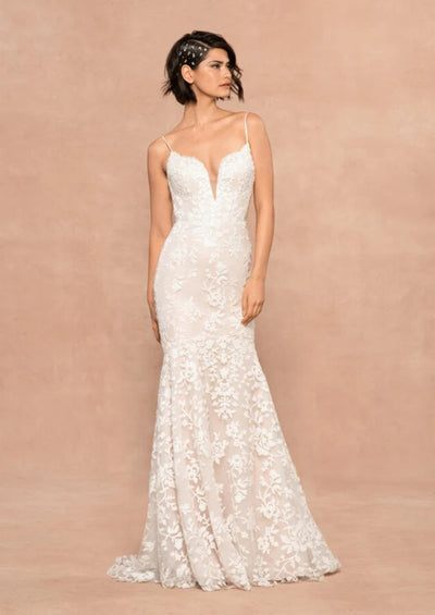 A woman wearing the Bergamot Bridal Hayley Paige Havana Mermaid/Trumpet Wedding Gown - Off The Rack, with a plunging sweetheart neckline and spaghetti straps, standing against a soft pink background.