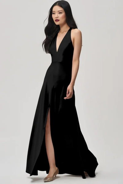 A woman stands elegantly in the Corinne Bridesmaid Dress by Jenny Yoo Off The Rack from Bergamot Bridal, featuring a sleek black satin back crepe material with a plunging halter V-neck and high slit, complemented by nude heels against a plain background.