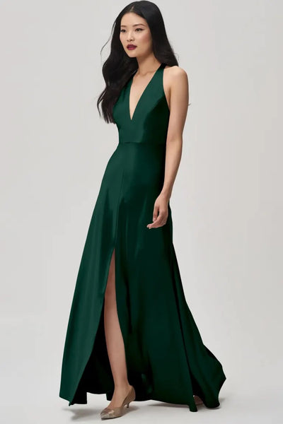 A woman wearing the Corinne Bridesmaid Dress by Jenny Yoo Off The Rack from Bergamot Bridal stands against a plain background. The deep green satin back crepe gown features a plunging halter V-neck and a high slit, perfectly complementing her long, dark hair and nude shoes, exuding effortless elegance.