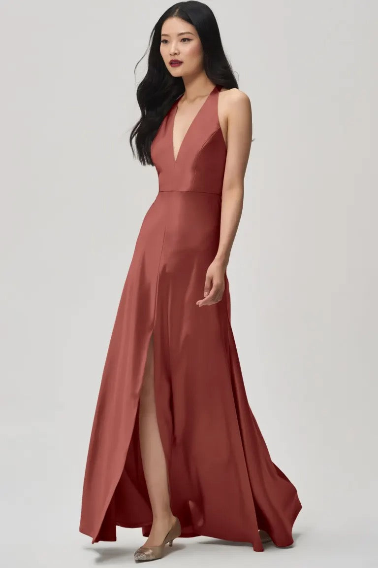 A woman poses elegantly in the Corinne - Bridesmaid Dress by Jenny Yoo Off The Rack from Bergamot Bridal, showcasing a sleeveless, floor-length bridesmaid dress in rust. It is crafted from satin-back crepe and features a plunging halter V-neck and a daring slit on the right leg.