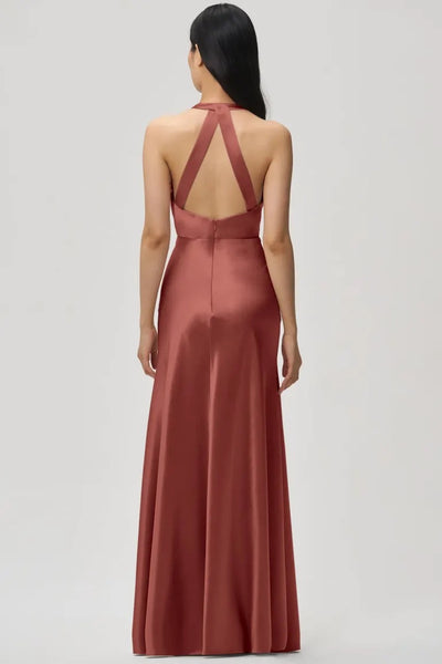 A woman in the Corinne - Bridesmaid Dress by Jenny Yoo Off The Rack from Bergamot Bridal, featuring a long, rose-colored satin back crepe fabric and elegant crisscross straps, is gracefully facing away.