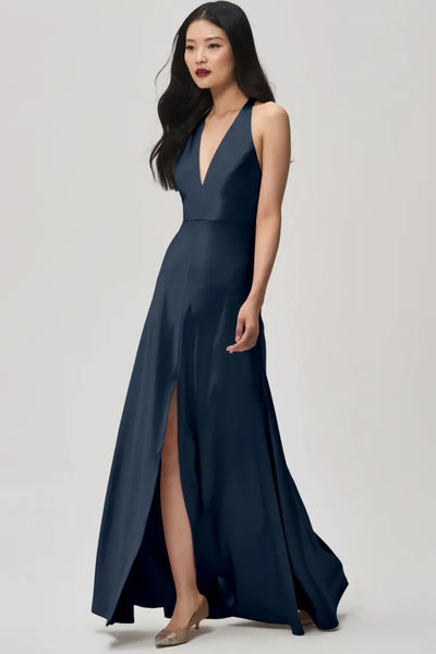 A woman in the Corinne bridesmaid dress by Jenny Yoo Off The Rack from Bergamot Bridal strides forward, highlighting its deep plunging halter V-neck and elegant slit against a plain background, crafted from satin back crepe fabric.