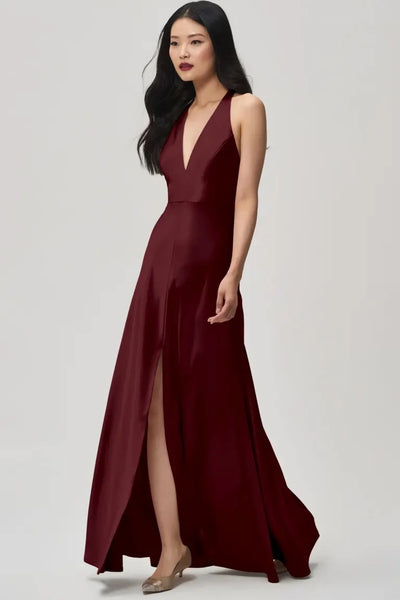 A woman in an elegant burgundy Corinne bridesmaid dress by Jenny Yoo satin back crepe with a plunging halter V-neck stands against a neutral background.