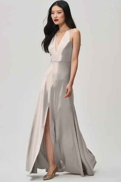 Gracefully walking against a plain background, a woman wears the Corinne bridesmaid dress by Jenny Yoo Off The Rack in silver-gray from Bergamot Bridal, featuring a plunging halter V-neck and high slit.