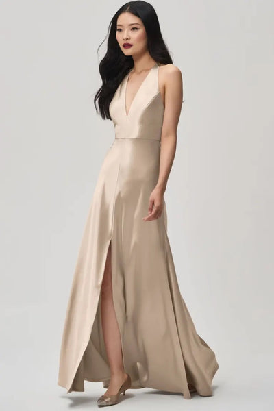 A woman in a long, Corinne Bridesmaid Dress by Jenny Yoo Off The Rack, with a beige satin back crepe material featuring a plunging halter V-neck and high slit, stands elegantly against a plain background.