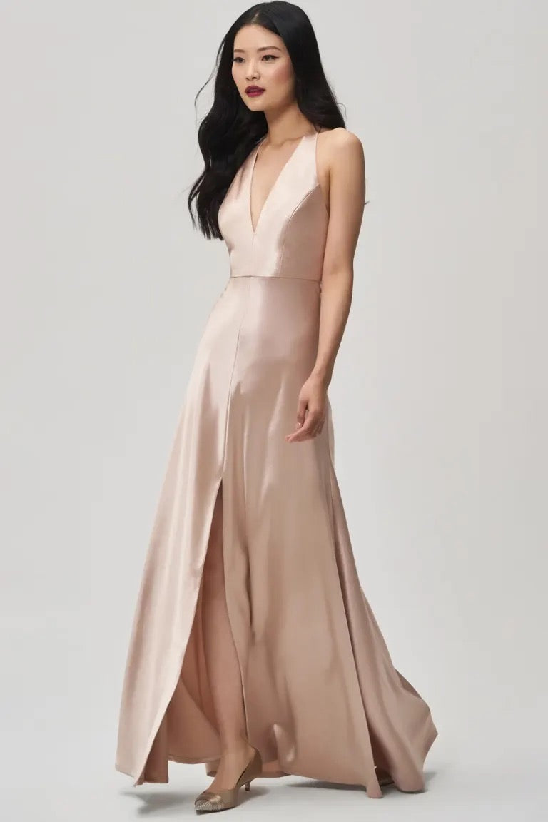 The woman stands elegantly against a simple backdrop wearing the Corinne bridesmaid dress by Jenny Yoo Off The Rack from Bergamot Bridal, a light pink, sleeveless gown crafted from satin back crepe with a plunging halter V-neck and high slit—an ideal selection for an elegant bridesmaid ensemble.