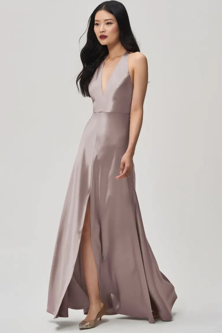 Clad in the Corinne Bridesmaid Dress by Jenny Yoo Off The Rack, a woman from Bergamot Bridal strides confidently against a plain backdrop, showcasing the sleeveless, floor-length design made of satin back crepe with a deep plunging halter V-neck and an elegant front slit.