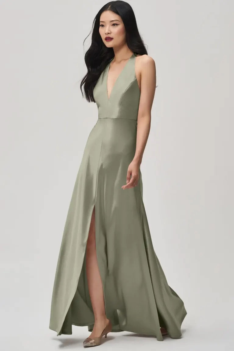 A woman in an elegant olive green satin back crepe Corinne - Bridesmaid Dress by Jenny Yoo with a plunging halter V-neck and a slit stands poised against a neutral background. (Brand Name: Bergamot Bridal)