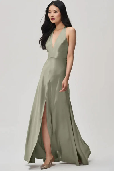 Wearing the Corinne bridesmaid dress by Bergamot Bridal, a woman elegantly walks forward in a satin back crepe fabric gown that boasts a sleeveless olive-green design and features a plunging halter V-neckline along with an effortlessly flowing front slit.