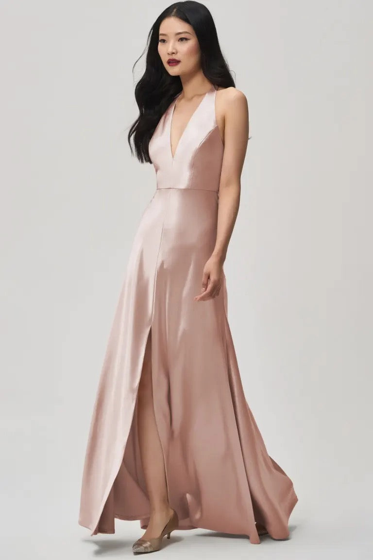 A woman wearing a sleeveless, plunging halter V-neck, blush-colored Corinne satin back crepe bridesmaid dress with an A-line skirt by Jenny Yoo at Bergamot Bridal.