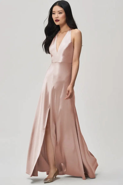 A woman wearing a sleeveless, plunging halter V-neck, blush-colored Corinne satin back crepe bridesmaid dress with an A-line skirt by Jenny Yoo at Bergamot Bridal.