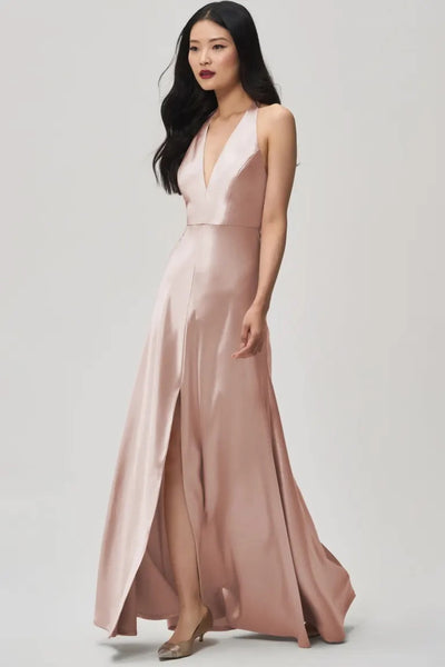 A woman stands against a plain background wearing the Corinne bridesmaid dress by Jenny Yoo Off The Rack, offered by Bergamot Bridal. This sleeveless gown in blush pink satin back crepe showcases a plunging halter V-neckline and a front slit, blending elegance with modern charm.