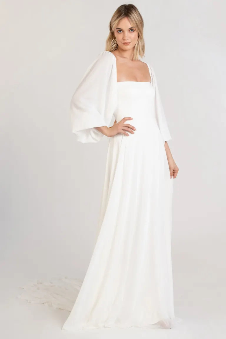 Woman in a Louise - Jenny Yoo Wedding Dress by Bergamot Bridal, with flowing chiffon sleeves, posing against a neutral background.