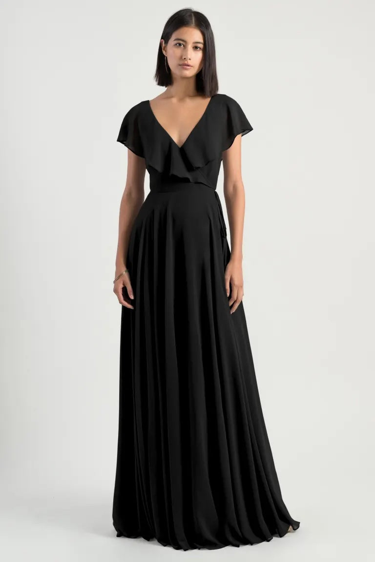 A woman in a black v-neck evening gown with flutter sleeves, the Faye - Bridesmaid Dress by Jenny Yoo, standing against a neutral background.
