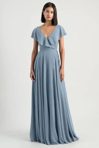 A woman in a pale blue, v-neck chiffon Faye bridesmaid dress by Jenny Yoo standing against a white background.
