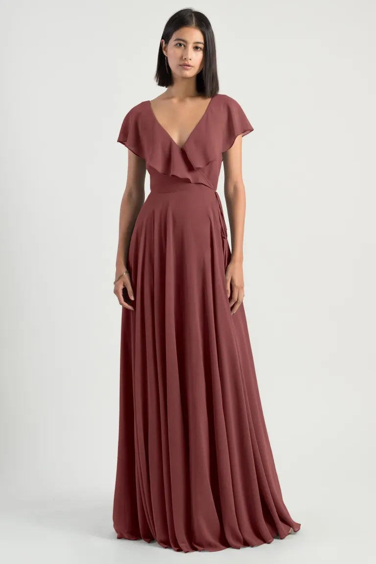 A woman in an elegant burgundy Faye bridesmaid dress by Jenny Yoo with flutter sleeves, standing against a neutral background from Bergamot Bridal.