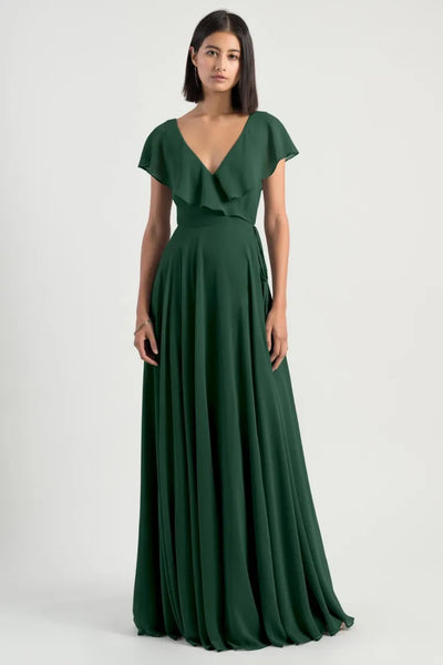 A woman in an elegant green chiffon evening gown with flutter sleeves and a v-neckline wearing the Faye - Bridesmaid Dress by Jenny Yoo from Bergamot Bridal.