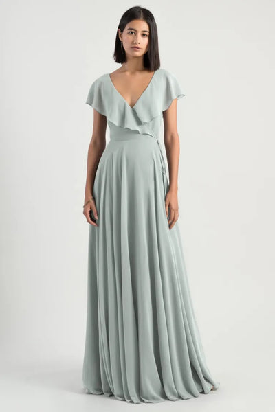 A woman posing in a long, flowing light gray chiffon Faye - Bridesmaid Dress by Jenny Yoo with flutter sleeves and a v-neckline from Bergamot Bridal.
