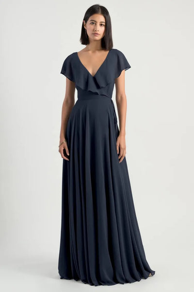Woman posing in an elegant navy blue Faye - Bridesmaid Dress by Jenny Yoo chiffon wrap dress with a V-neckline, flutter sleeve, and flowing skirt from Bergamot Bridal.